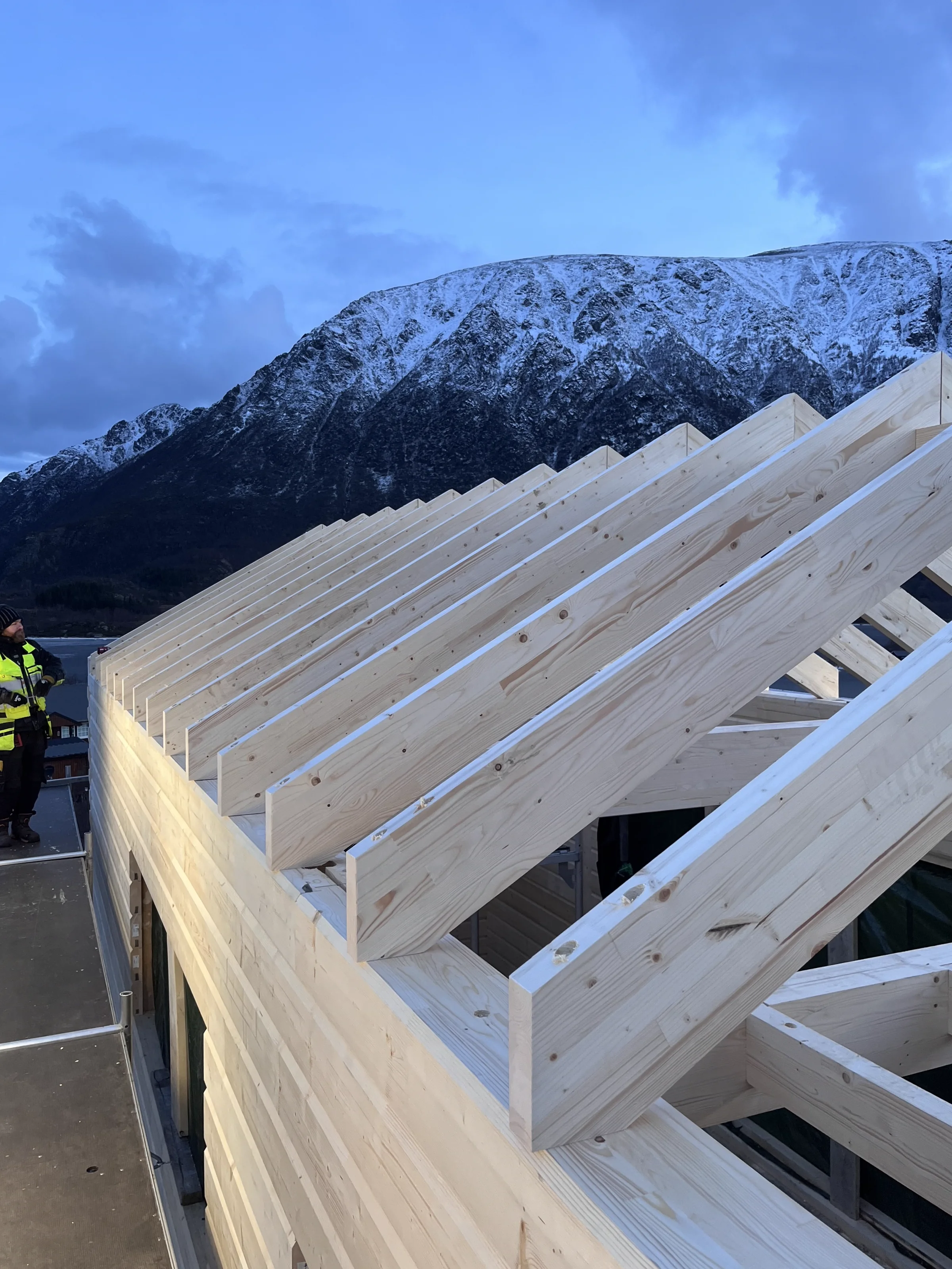 Aufgerichteter Dachstuhl aus hellem Holz vor schneebedecktem Bergpanorama in der Abenddämmerung